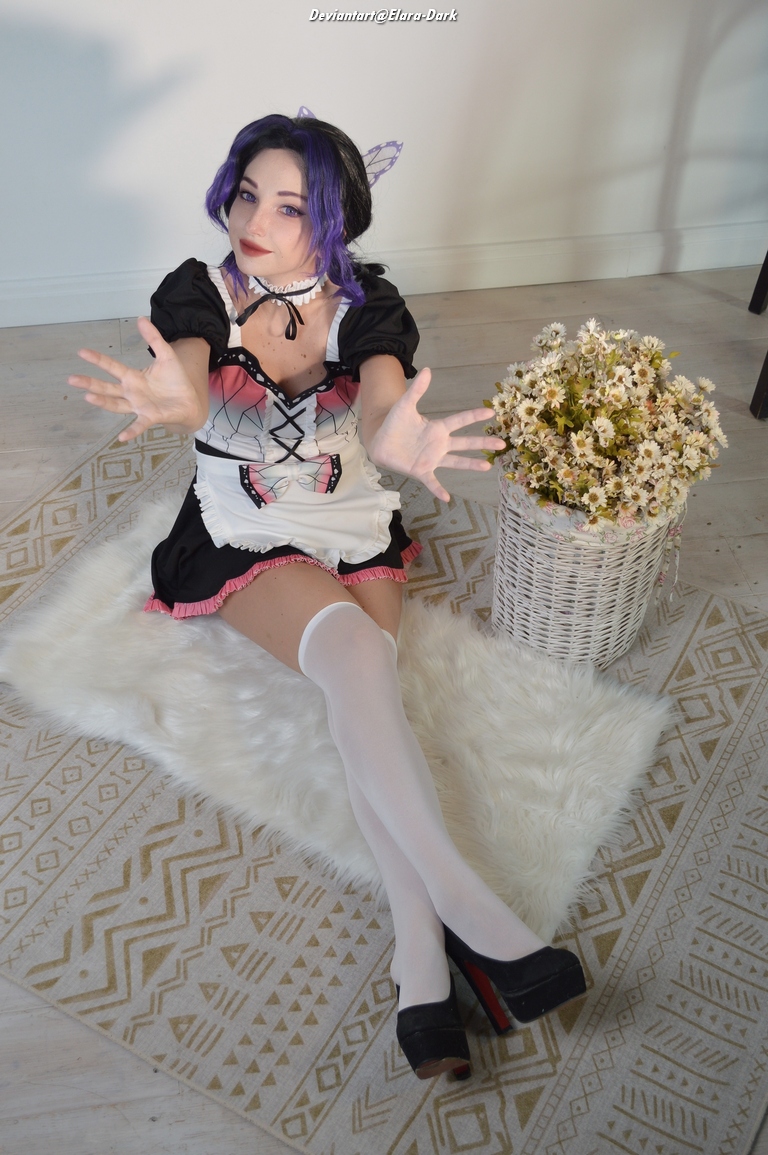A person wearing a black and pink dress with white stockings and high heels sits on a fluffy rug, posing with outstretched hands, surrounded by a decorative basket of flowers.