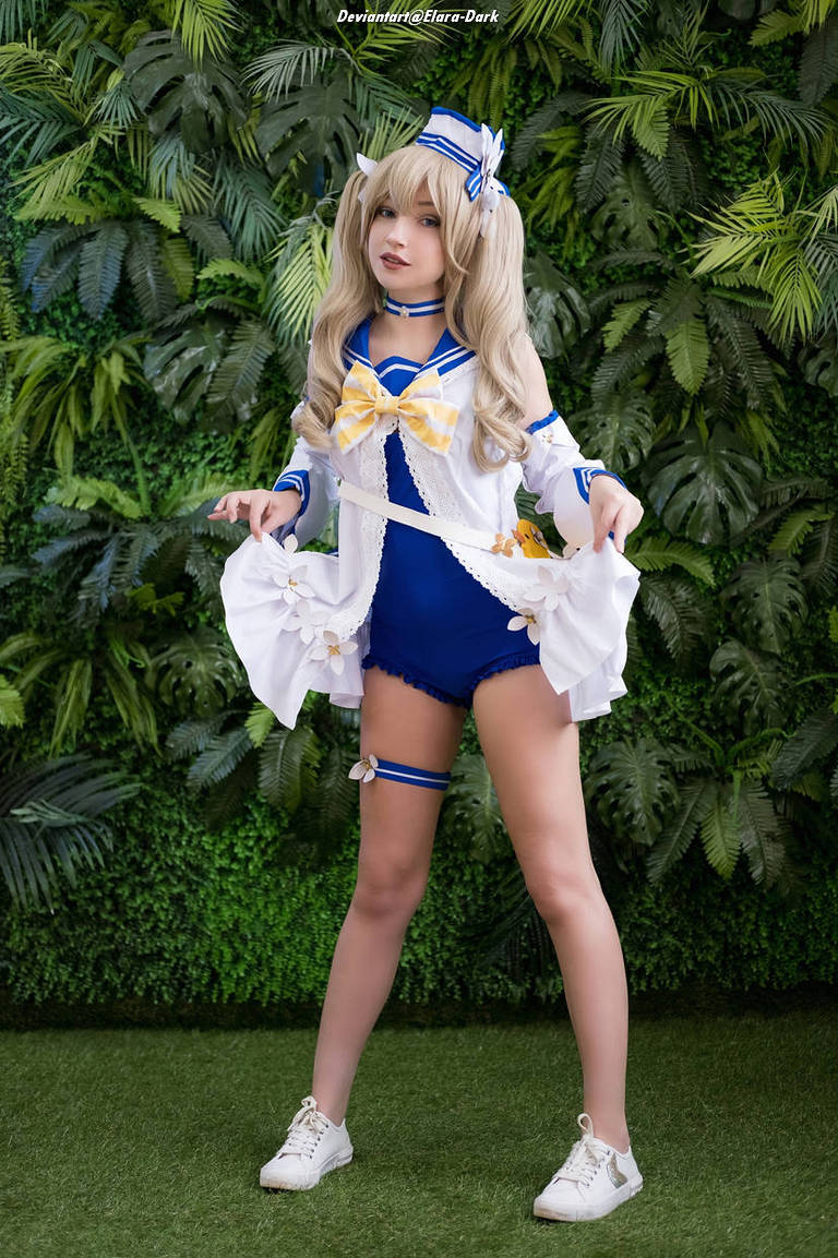 A person in a blue and white cosplay outfit with a bow, posed in front of a leafy green backdrop.
