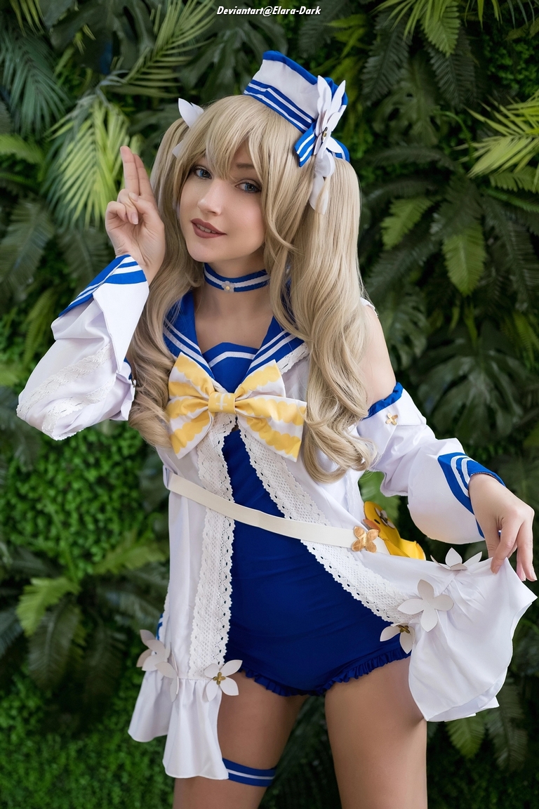 A person in a colorful costume with a blue and white theme, featuring a large bow, poses with a playful hand gesture in front of lush greenery.