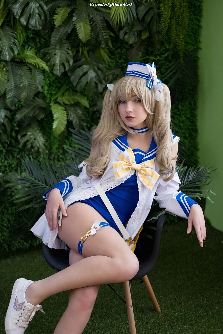 A person with long blonde hair sitting elegantly in a chair, wearing a blue and white sailor-style outfit with a large bow. The background features green foliage, adding to the vibrant atmosphere.