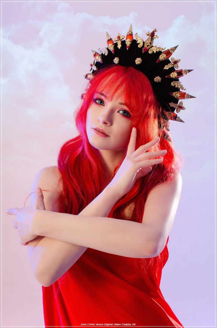 A young woman with long red hair poses gracefully, wearing a striking black crown adorned with spikes and gold accents. She is dressed in a vibrant red outfit, with a soft pastel background.