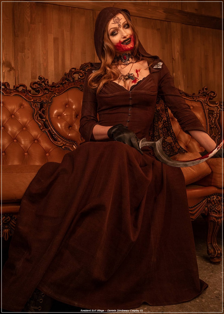 A woman dressed in a dark brown hooded gown with intricate details sits on an ornate leather couch, holding a curved blade. Her makeup features dark accents and a blood-like design, creating a striking and eerie appearance.