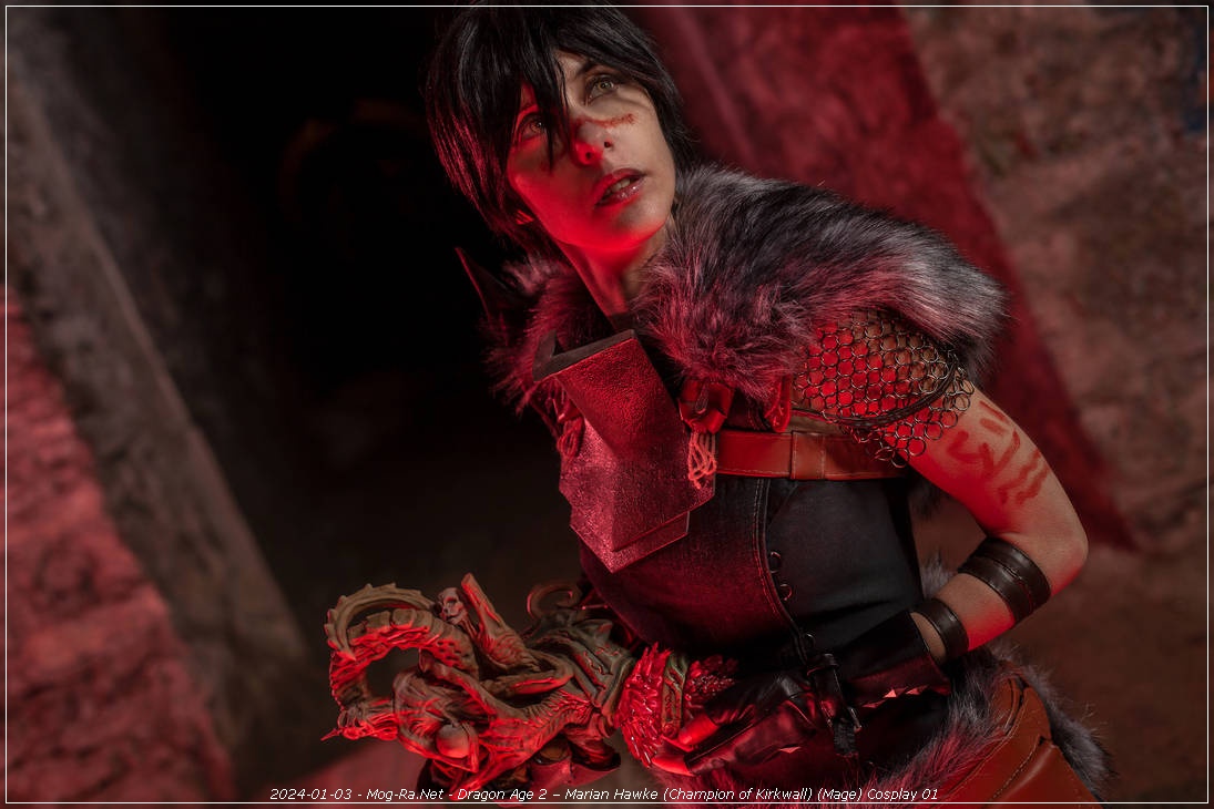 A person in a fantasy costume, representing a character from a video game, stands in a dimly lit environment. They have short black hair, facial markings, and wear a dark outfit with fur accents and intricate accessories, including a decorative staff.