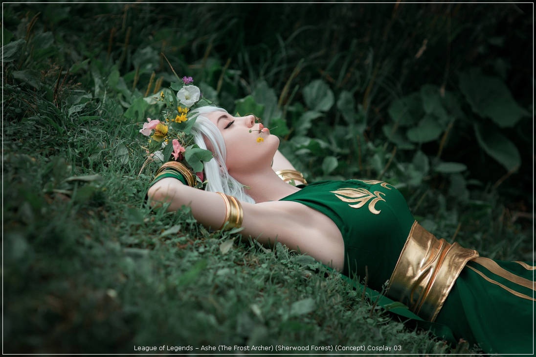 A cosplayer dressed as Ashe from League of Legends, lying in a lush green setting with flowers in her hair and a serene expression.