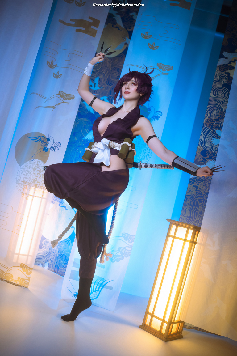 A cosplayer dressed in a dark outfit strikes a dynamic pose, holding kunai knives, with a backdrop featuring blue and gold decorative fabric and two illuminated lanterns.