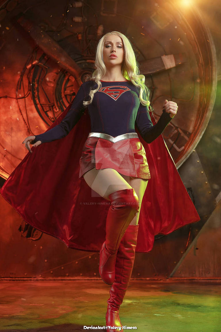 A female superhero model wearing a blue and red costume, featuring a logo on the chest, long blonde hair, and a red cape, posed dramatically against a colorful, illuminated background.