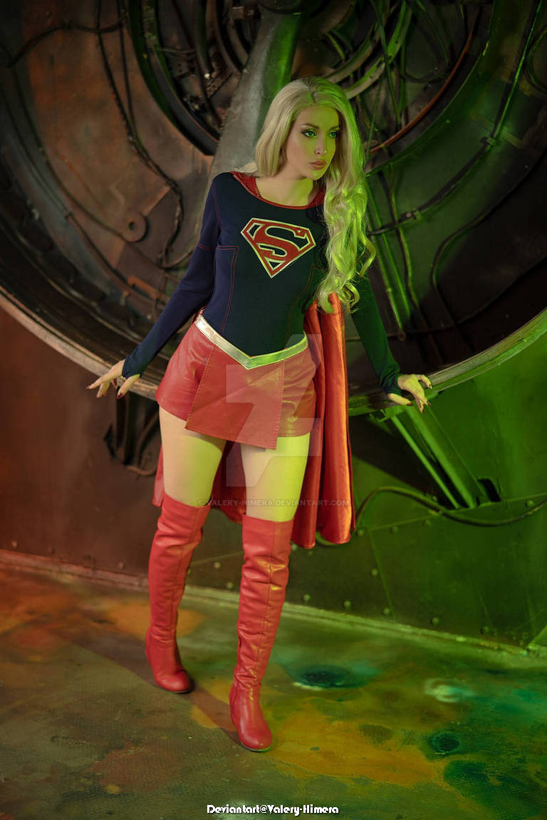 A person dressed as a superhero with long blonde hair stands in a striking pose, wearing a blue and red costume with a cape and high red boots. The background features mechanical elements with green lighting.