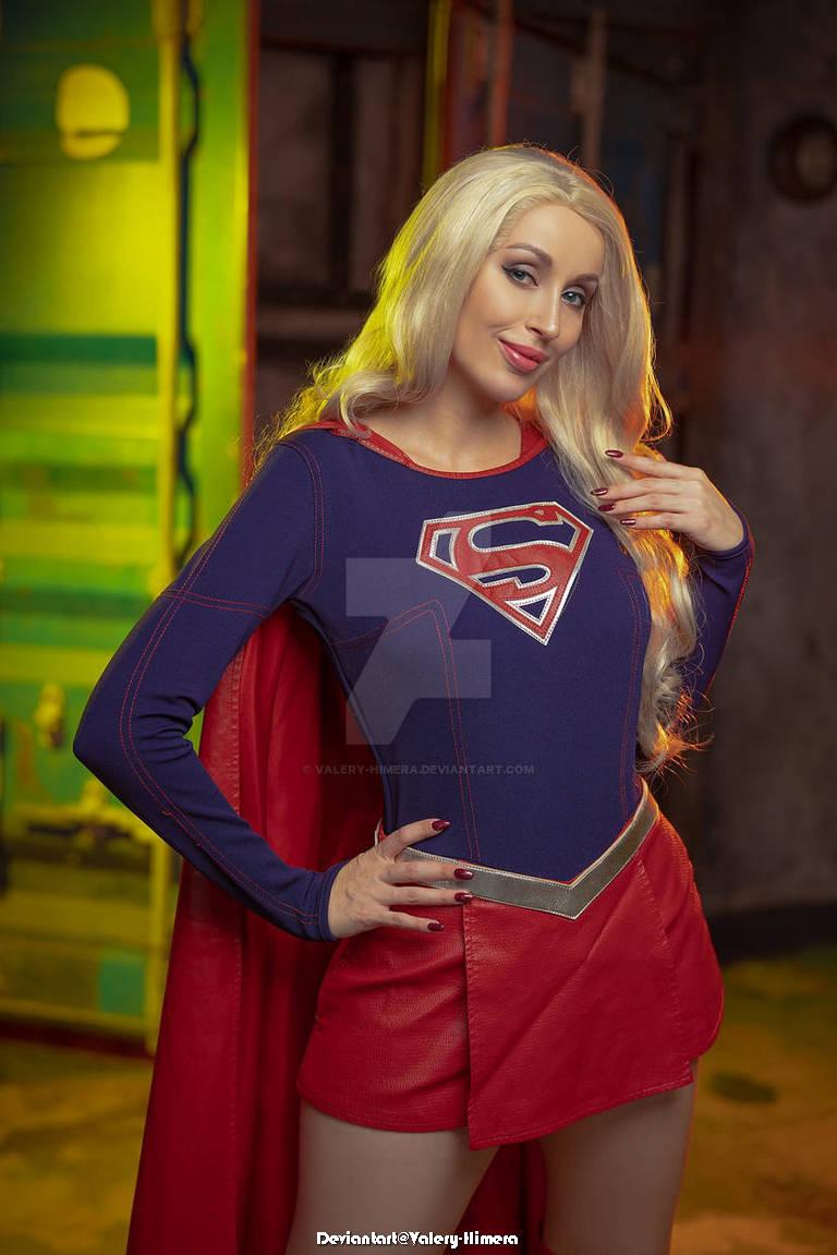 Woman dressed as a superhero with a red cape and blue outfit featuring an emblem on the chest, posing confidently with a smile in a colorful studio backdrop.