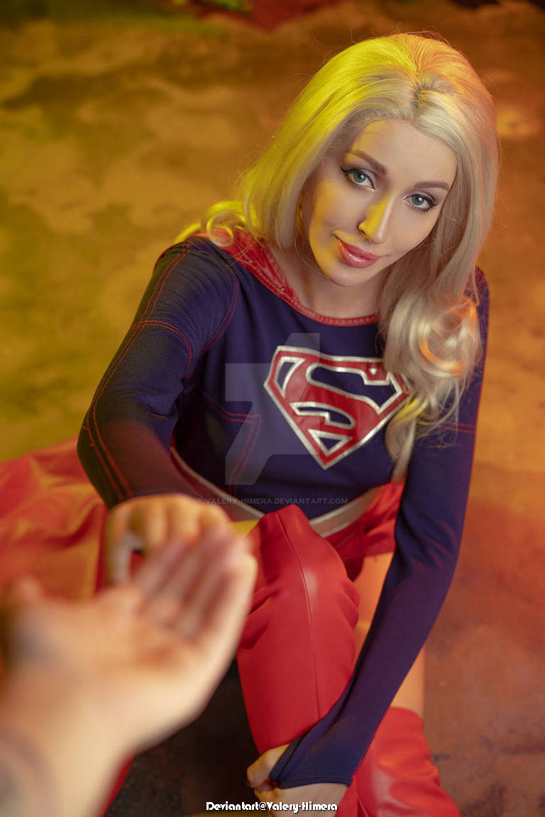 A character dressed as Supergirl, with long blonde hair, sitting on the floor and extending a hand towards the viewer, creating a warm and inviting atmosphere.