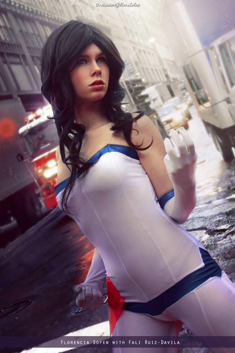 A young woman dressed as a superhero, wearing a white and blue costume with a red cape, poses confidently on a city street.