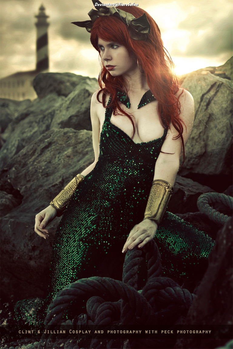 A woman with long red hair wearing a green sequined dress sits on rocks by the coastline, with a lighthouse in the background.