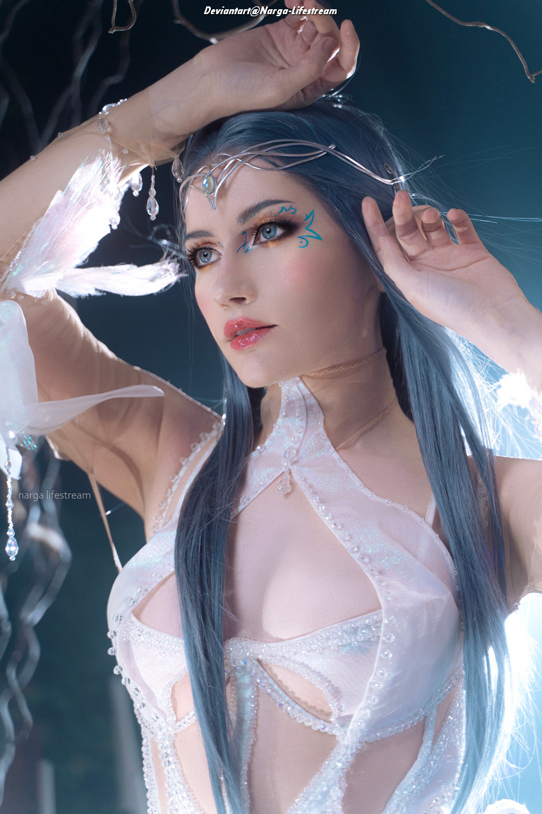 A close-up portrait of a female figure with long blue hair and elaborate makeup, wearing a sheer, intricately designed dress embellished with beads and translucent details.