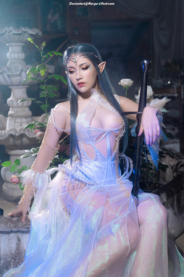 A character dressed in an ethereal, flowing gown with intricate patterns, seated gracefully. She has long, flowing blue hair and pointed ears, holding a dark staff beside her, surrounded by soft, ambient lighting and floral elements.