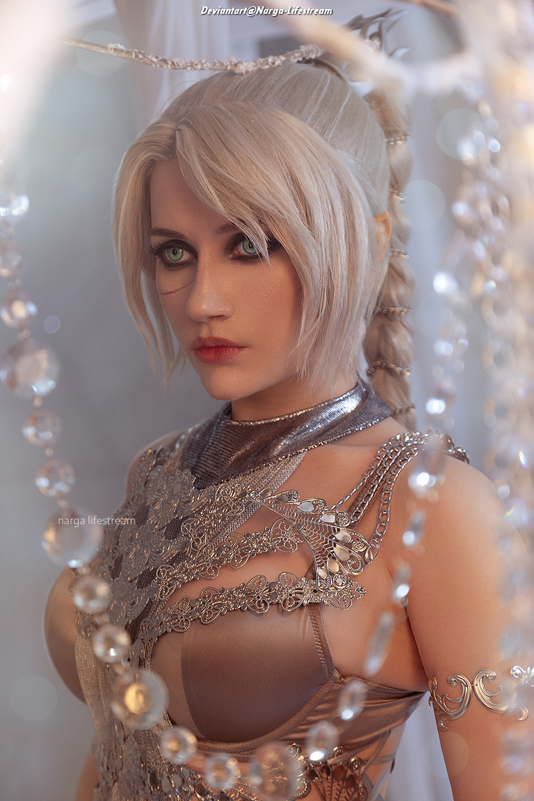 A close-up portrait of a person with light blonde hair styled elegantly, wearing intricate metallic jewelry and a detailed outfit. The background features sparkling beads and a soft focus, enhancing the ethereal vibe of the image.