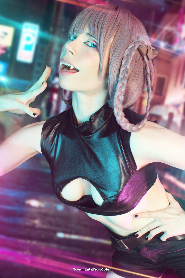 A person with short, light purple hair wearing a black outfit poses dramatically with an exaggerated expression, showcasing fang-like teeth against a colorful city background.