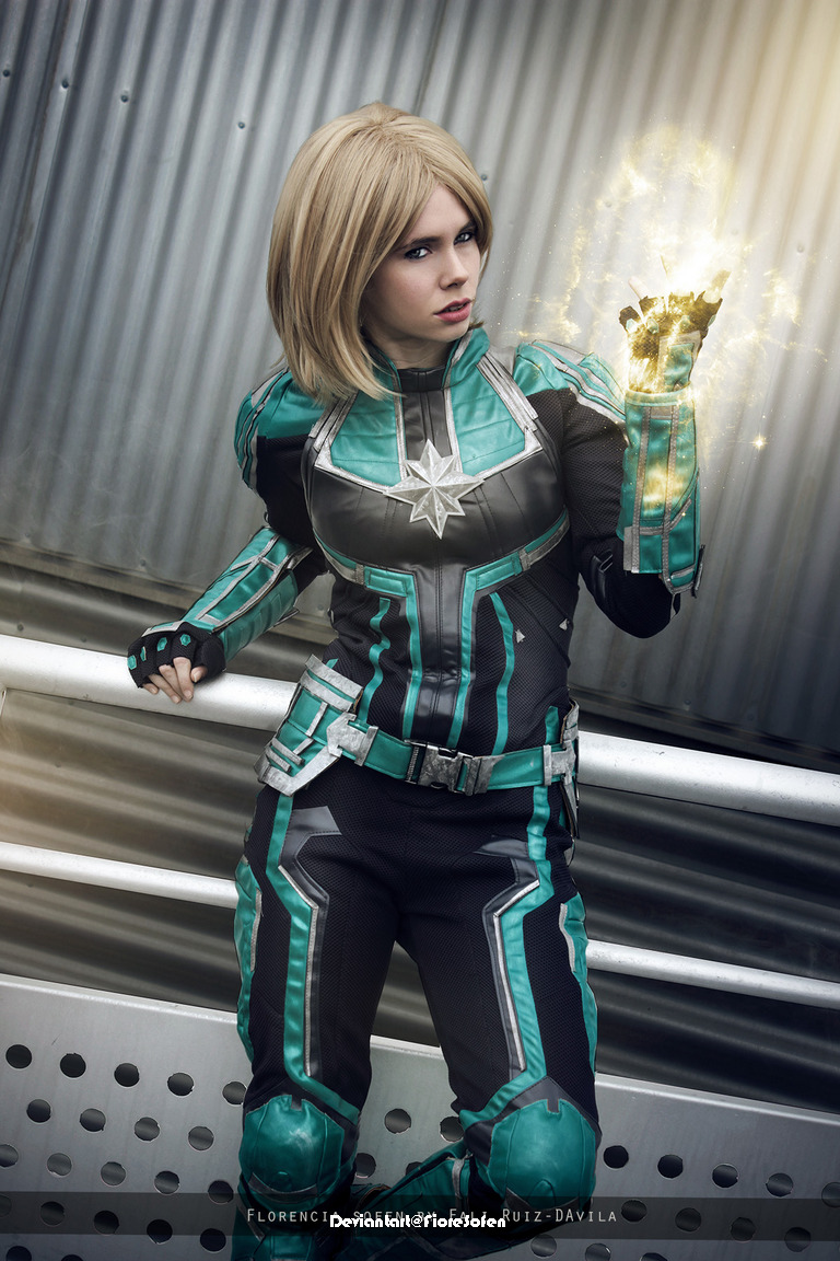 A person in a superhero costume with turquoise and black details is displaying energy emanating from their hand, set against a metallic backdrop.