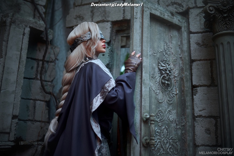 A person in a dark cloak with a braided hairstyle and a decorative headpiece stands beside an ornate door, appearing to look into an old, mysterious building.