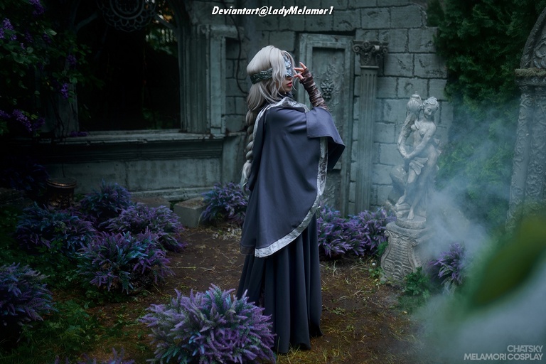 A figure in a flowing gray costume with silver hair and intricate accessories stands in a mystical garden filled with purple plants, facing away from the camera. The background features stone structures and fog, creating a magical atmosphere.