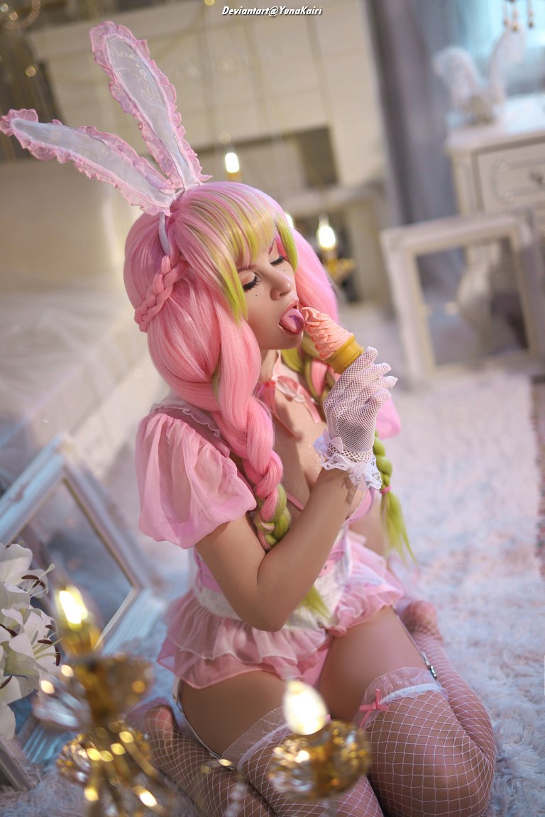 A person with pink hair and bunny ears wearing a pink outfit is licking an ice cream cone while sitting on a soft surface decorated with candles.