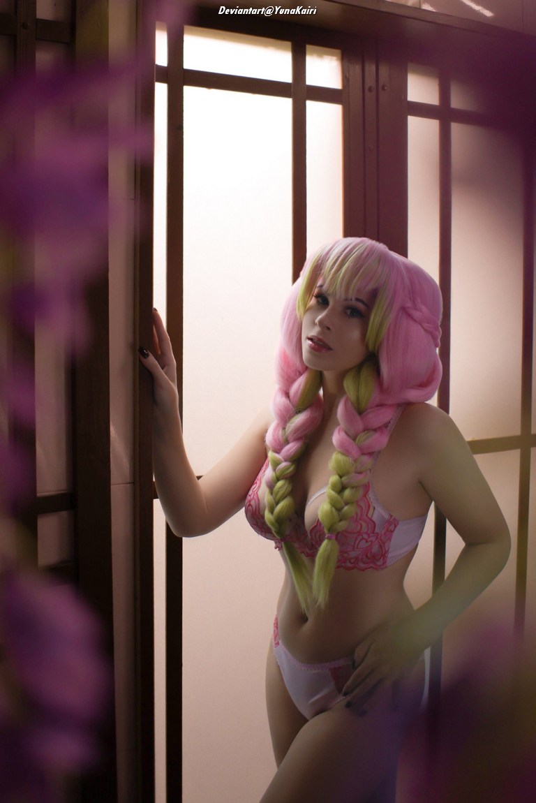 A model with long, pink and green braided hair poses elegantly by a wooden screen, wearing intricate lingerie with lace details and a soft expression.