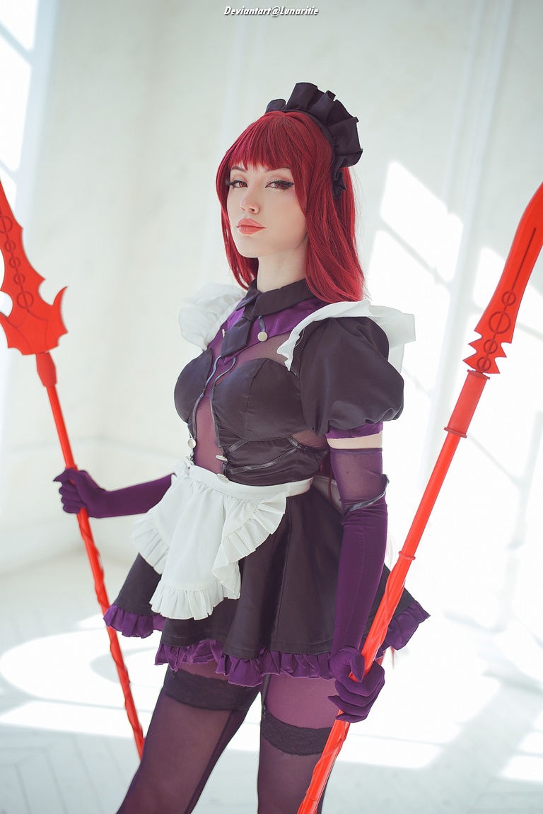 Cosplayer dressed in a black and purple maid outfit, holding two red staff-like weapons, with long red hair and dramatic makeup, posed in a well-lit indoor setting.