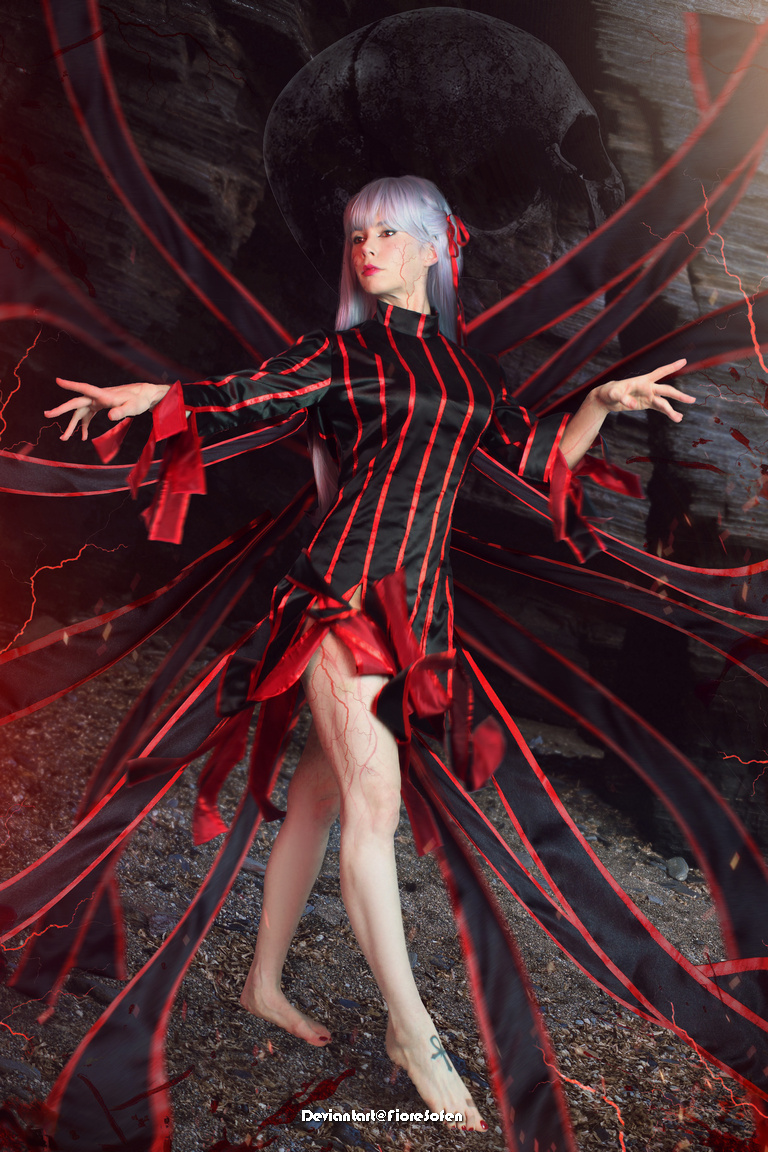 A person in a striking black and red outfit with dramatic sleeves, posing confidently against a dark, atmospheric background with a skull and red accents.