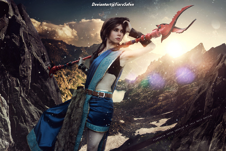 A person dressed in a blue and black costume, holding a red weapon, poses dramatically against a mountainous landscape with a dramatic sky in the background.