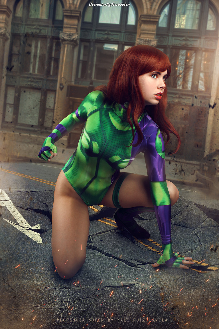 A woman with long red hair wearing a green and purple bodysuit poses in a dynamic crouching position on a cracked street with a background of dilapidated architecture.