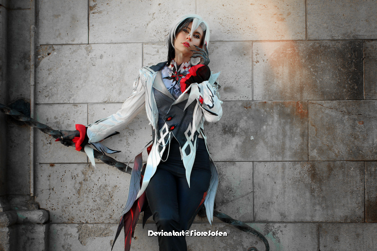 A person in a detailed costume inspired by a character, leaning against a textured stone wall. The outfit features a mix of colors, elegant patterns, and accessories, conveying a dramatic and artistic appearance.