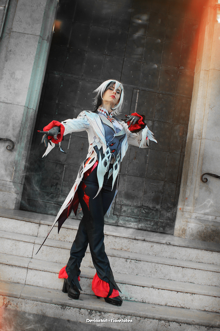 A person posing in a detailed costume with white and red elements, standing on stone steps in front of a dark doorway.