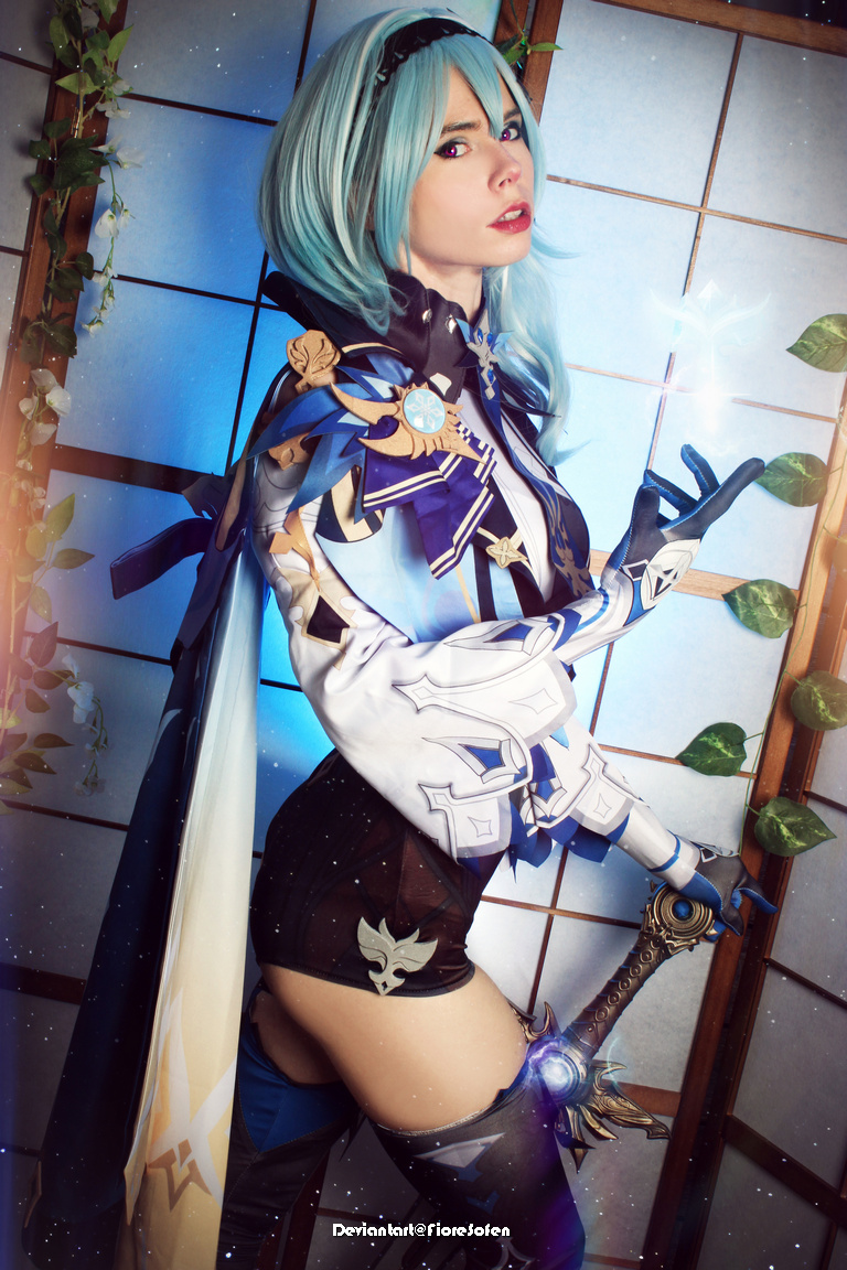 A person dressed in a detailed blue and white costume with fantasy elements, posing in front of a Japanese-style screen. Their hair is blue, and they are holding a glowing object.