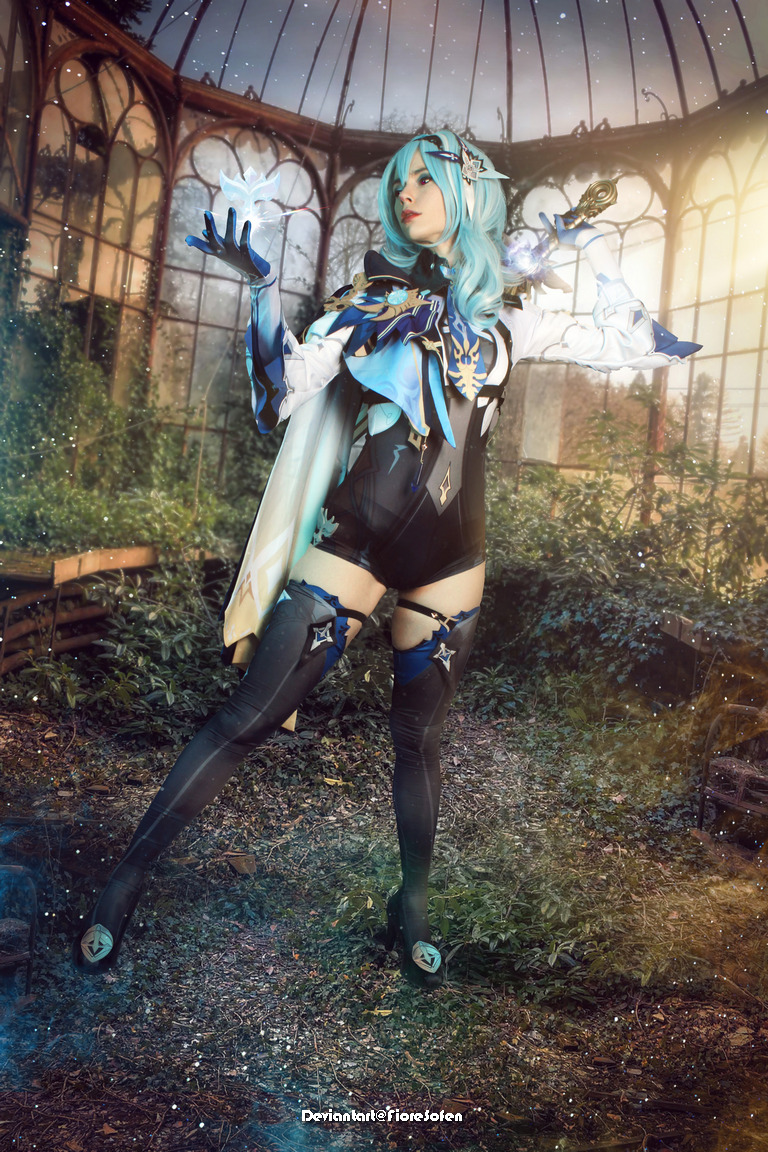 A person in a fantasy-themed costume with blue hair poses dramatically in a lush, overgrown glass greenhouse. They appear to be casting magic with glowing elements in their hands.
