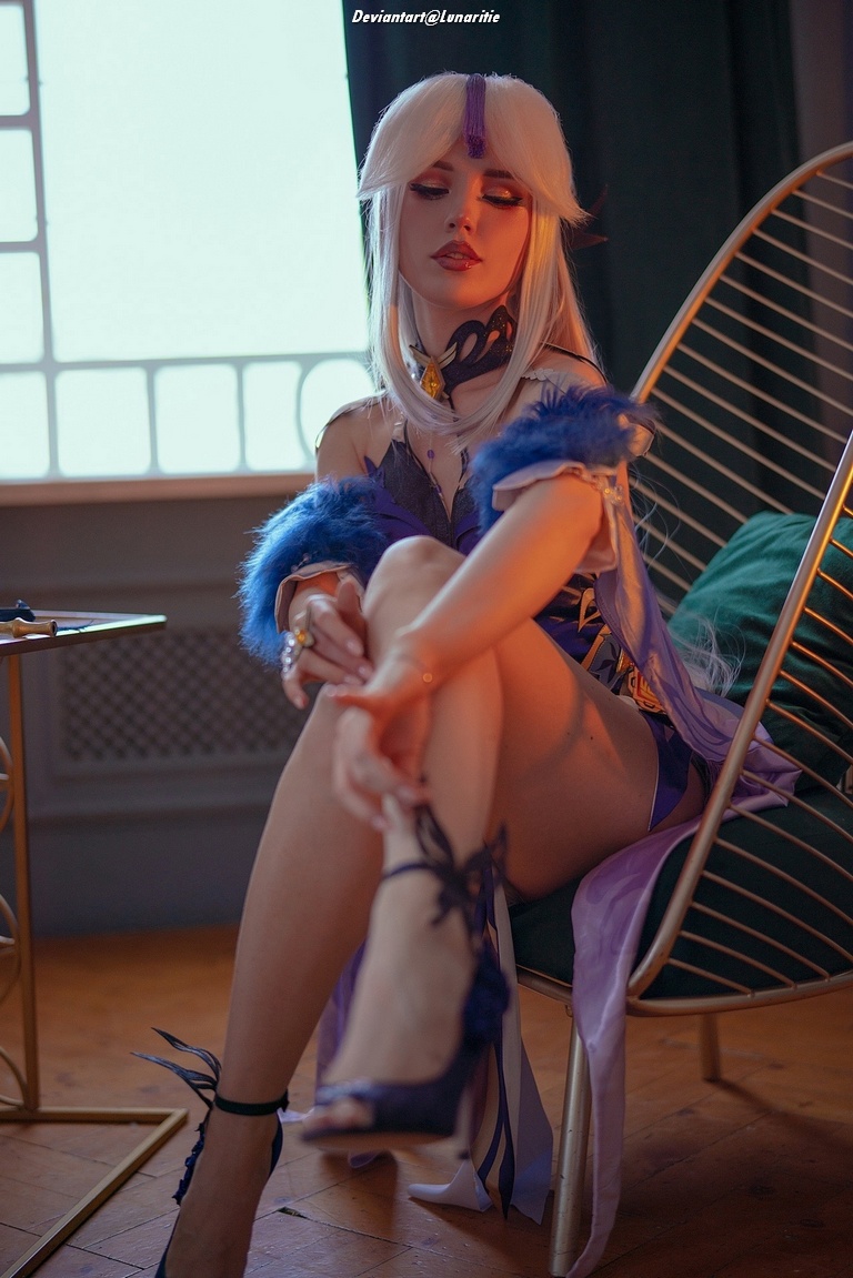 A person dressed in an elaborate costume with light purple tones and fur accents sits in a stylish chair, showcasing a thoughtful expression and emphasizing intricate footwear.