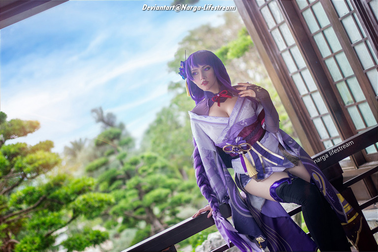A woman in a purple and white costume stands on a balcony, surrounded by lush greenery and traditional architecture, with a serene expression.