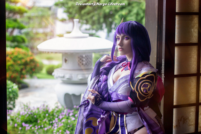 A person in a fantasy costume with purple hair and elaborate clothing stands by a window, gazing outside into a serene garden setting.