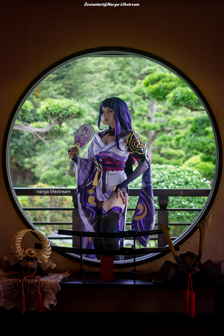 A person in a traditional costume stands by a circular window, holding a fan, with a lush green garden visible outside.