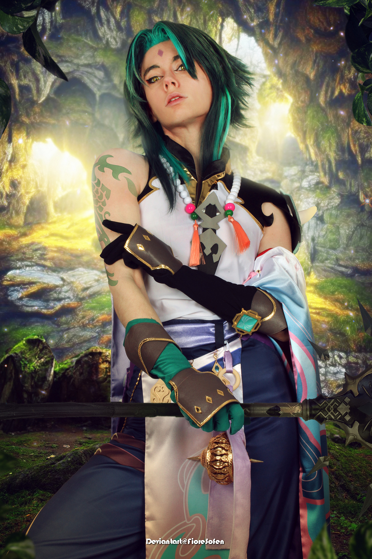A person in a fantasy-themed costume with green hair, striking a confident pose. The background features a mystical forest with soft lighting and natural elements.