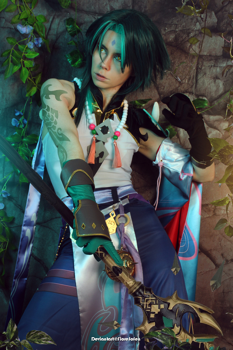 A person dressed in an elaborate costume, featuring teal hair and intricate accessories, poses with a sword against a backdrop of rocks and foliage.