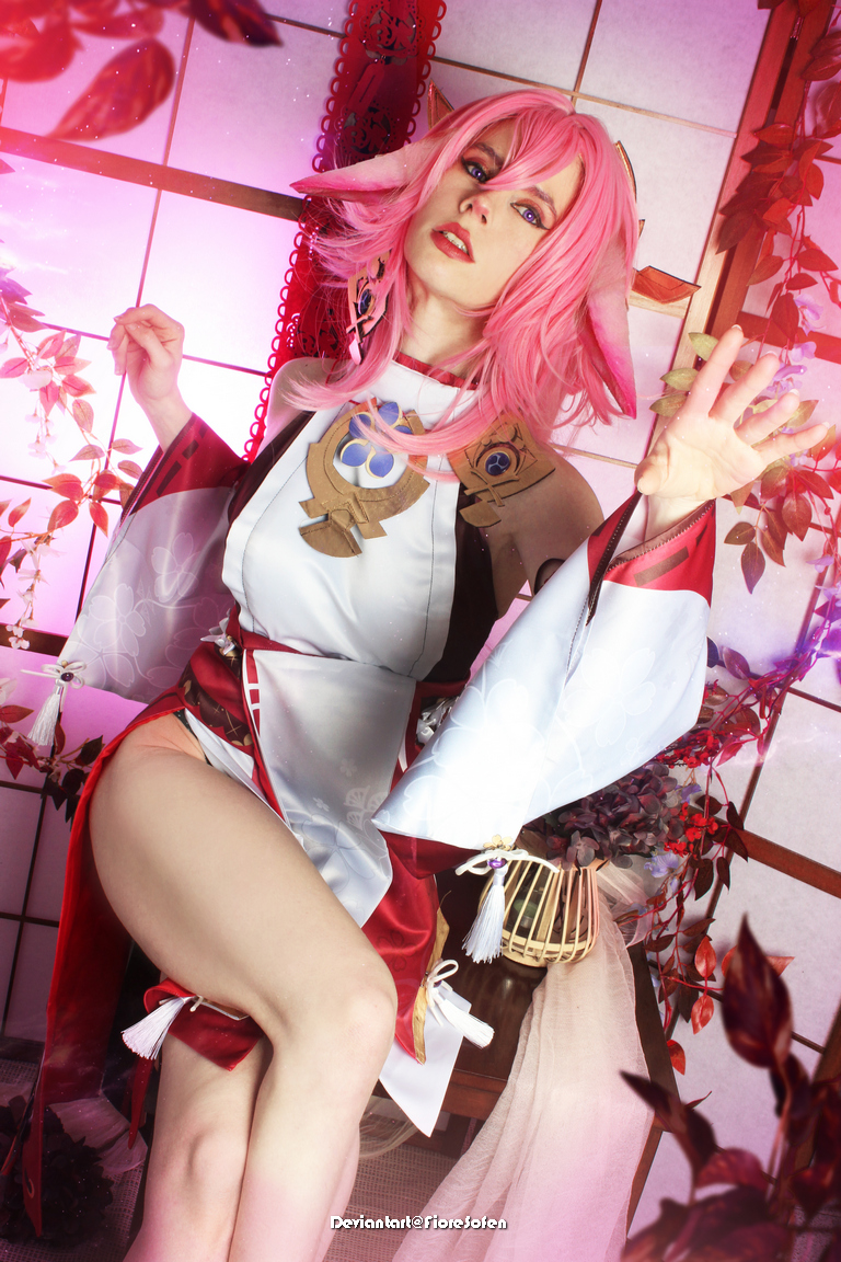 A person wearing a colorful fantasy costume with pink hair and elaborate accessories, posed dramatically among decorative plants and backdrops.
