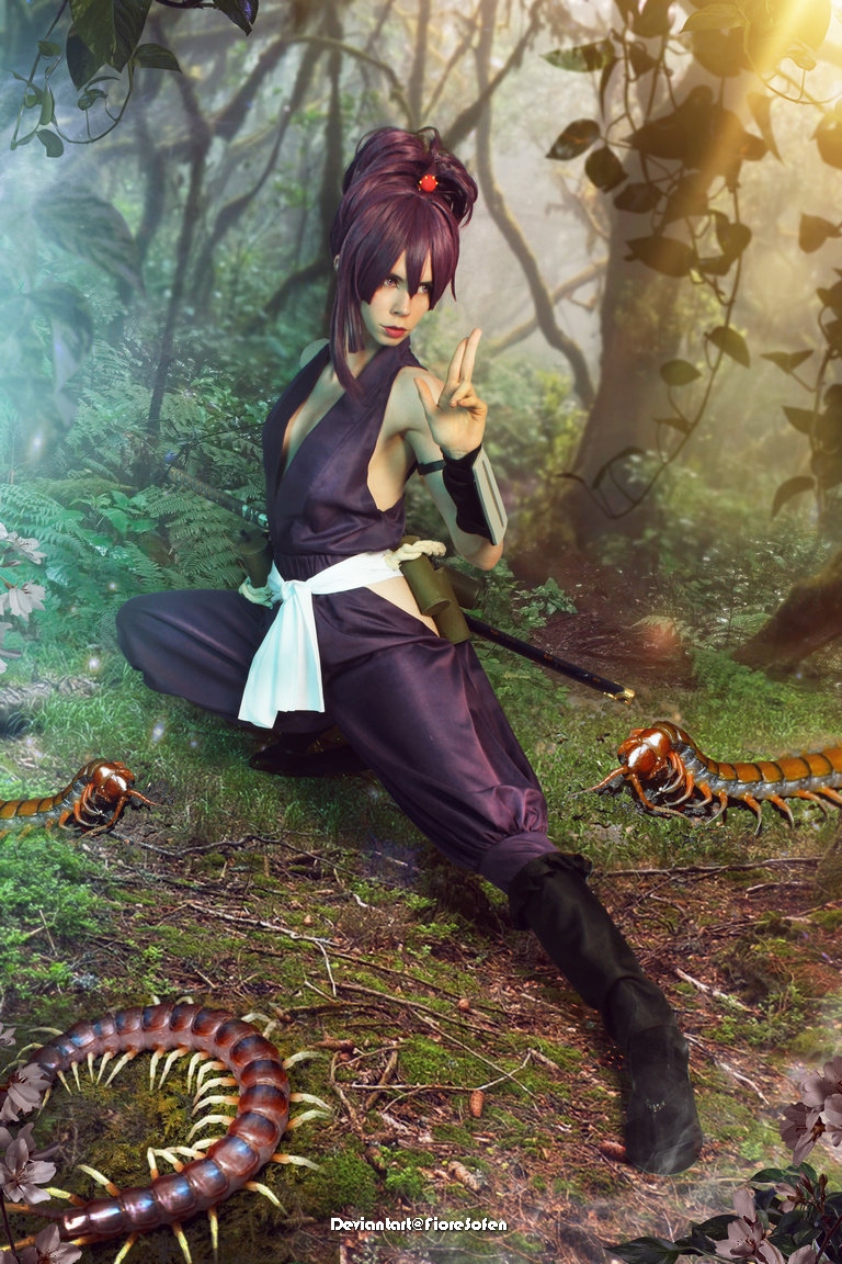 A female cosplayer dressed in a dark outfit poses in a forest, surrounded by large centipedes, with a confident expression and a peace sign gesture.