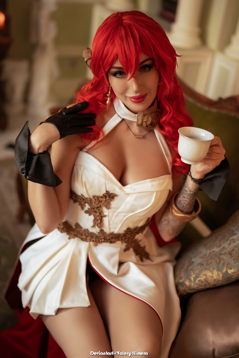 A woman with vibrant red hair wearing an elegant white dress with gold details, sitting on a decorative chair, holding a teacup and smiling.
