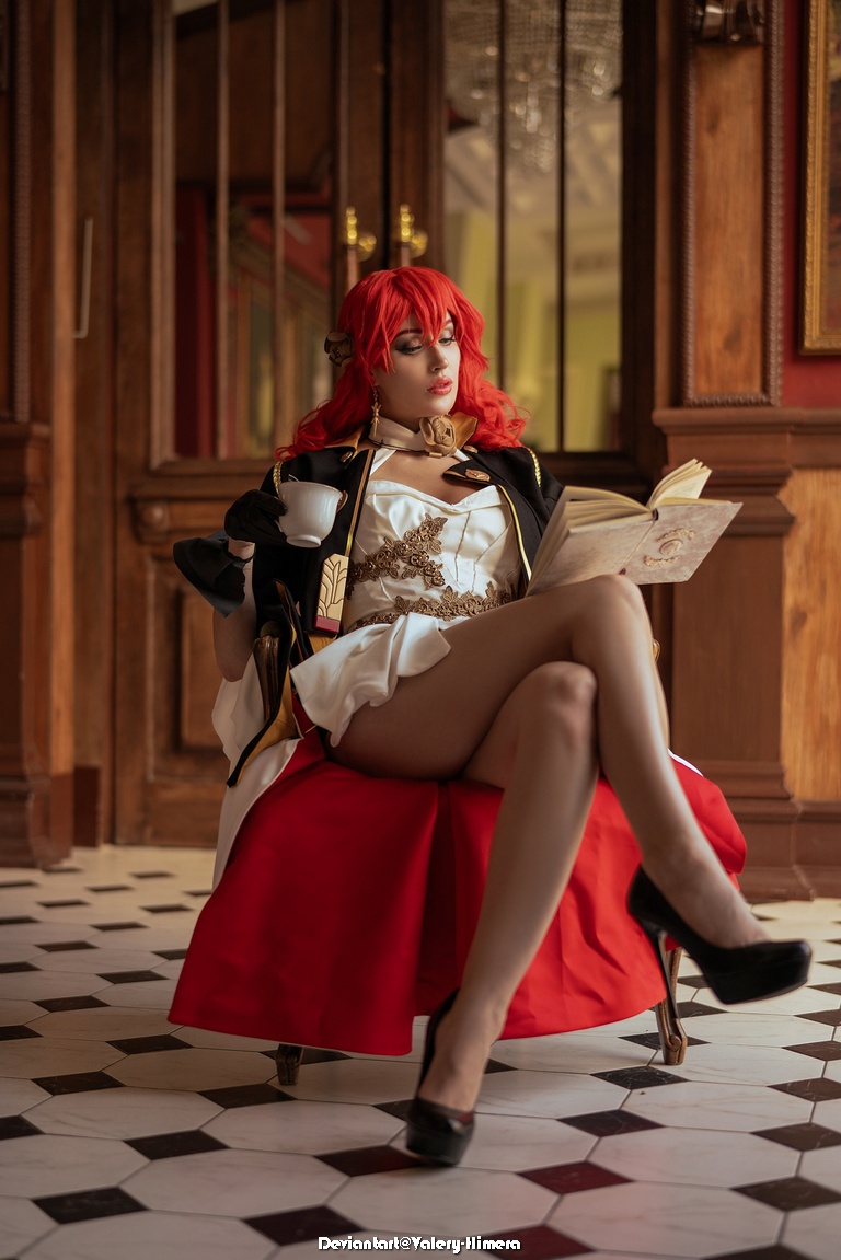 A person with red hair sits in a chair wearing a stylish outfit, holding a cup and reading a book, in an elegantly decorated interior.