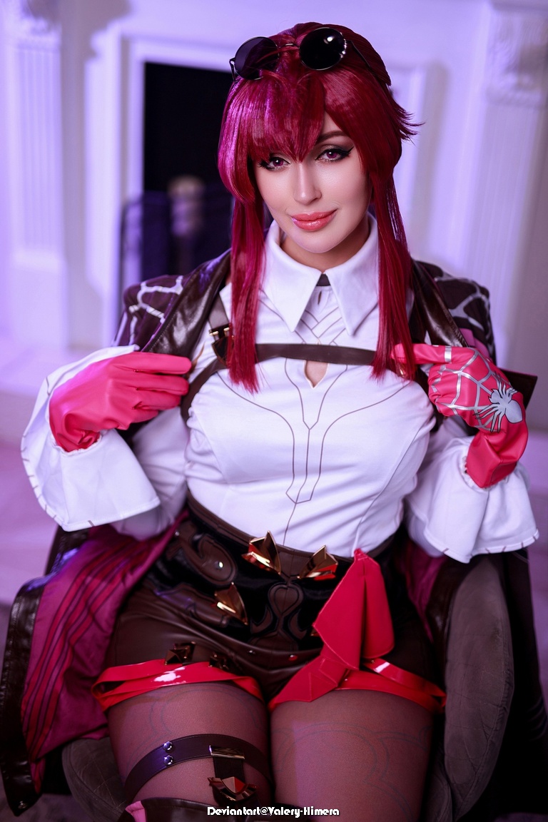 A person cosplaying in a stylish outfit with a white shirt, a brown jacket, and pink accessories, sitting in a chair with a playful expression.