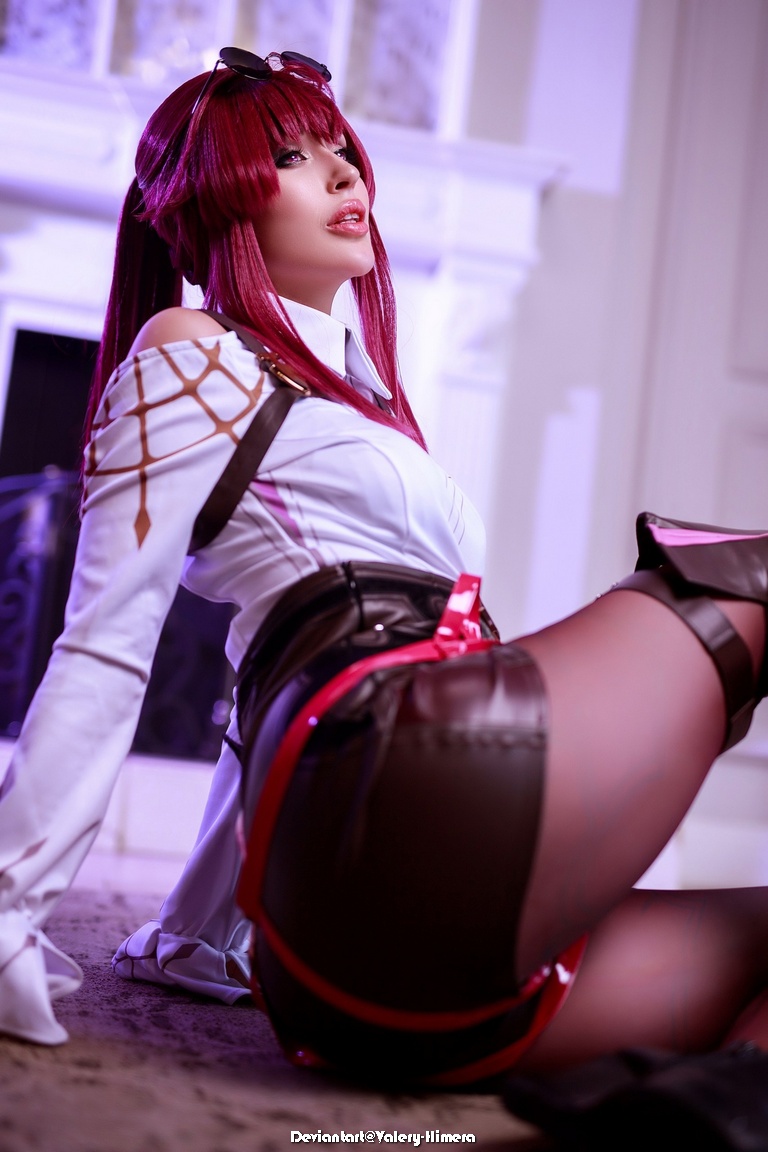 A person in cosplay with long red hair, wearing a white and black outfit, poses on the floor in a stylish manner.