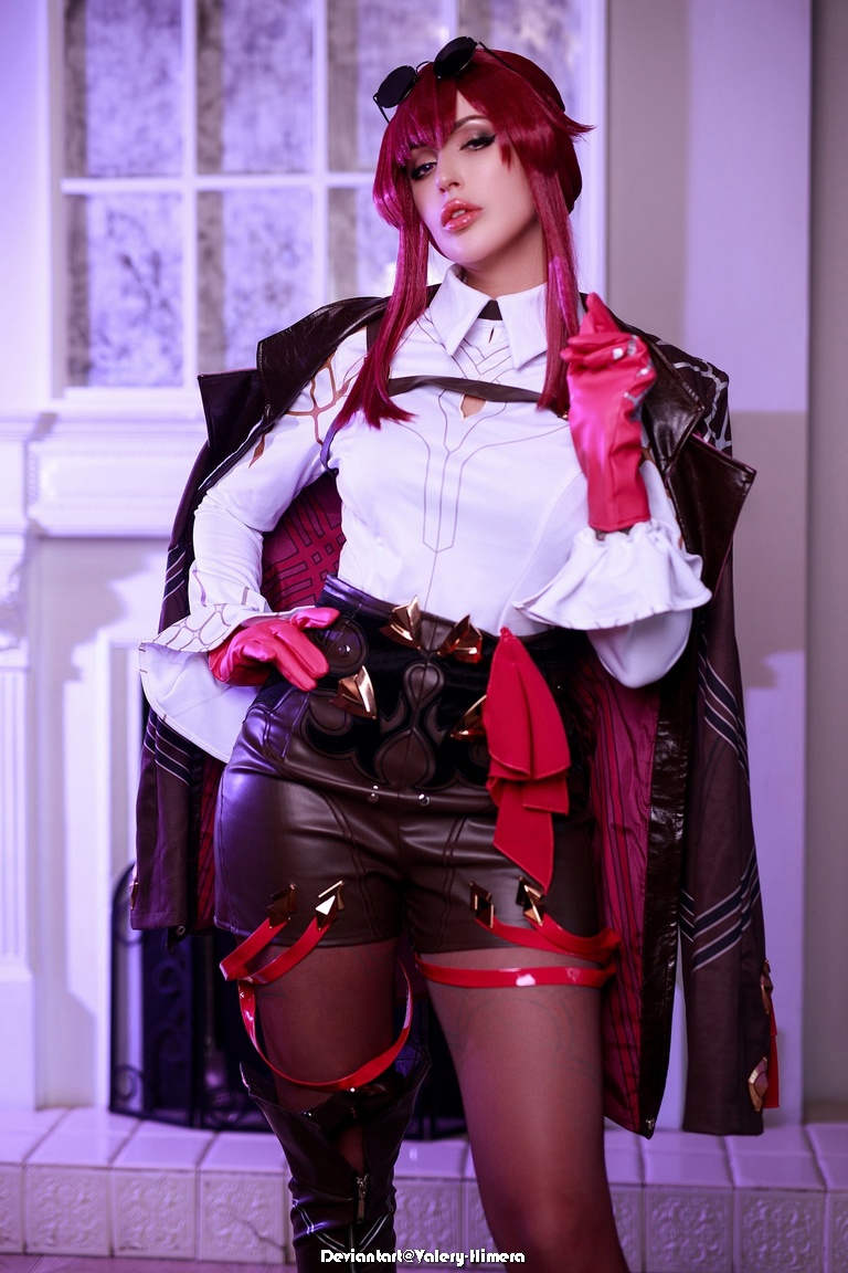 A person in a detailed costume, featuring a white shirt, leather shorts, and a long dark coat, with red gloves and accessories. The character has long red hair and is posed in a well-lit indoor setting.
