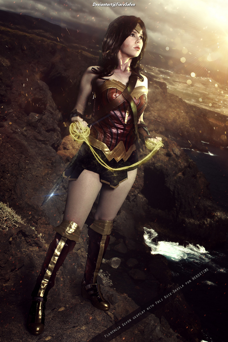A female cosplayer dressed as Wonder Woman stands confidently on rocky terrain, holding a lasso in her hands. The background features a dramatic sky and ocean waves.