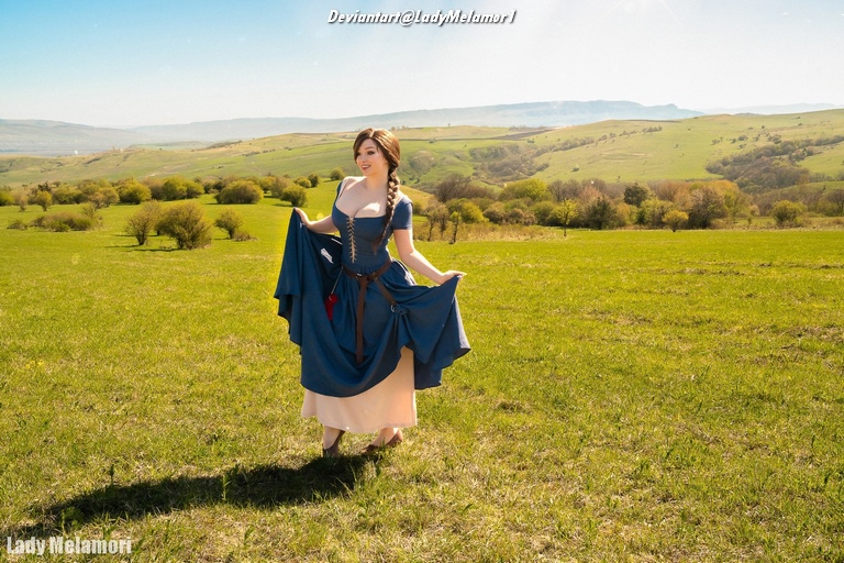 A woman in a historical dress standing in a green field with rolling hills in the background.