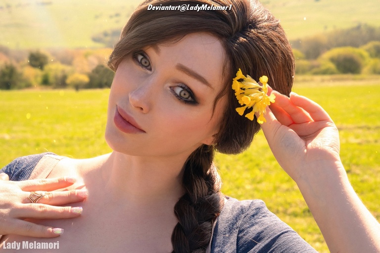A close-up portrait of a woman with long, braided hair, holding a yellow flower next to her face, set against a green landscape.