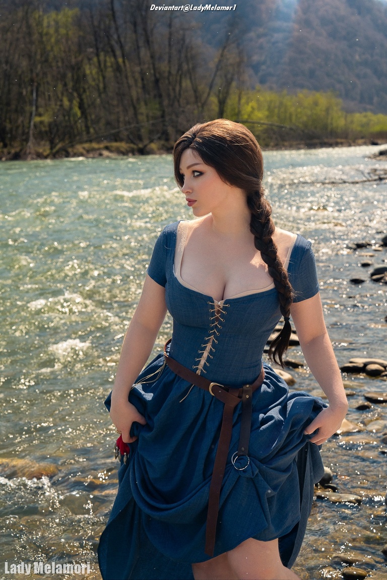 A woman in a blue dress standing by a river, with long hair and a braid, surrounded by trees and mountains.