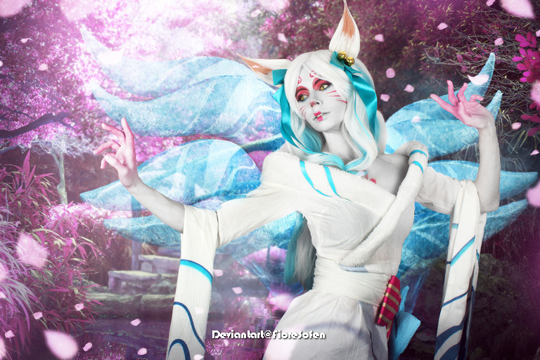 A character with fox-like features wearing a white robe and blue accents, surrounded by flowering cherry blossom trees, with ethereal, flowing blue tails and a serene expression.